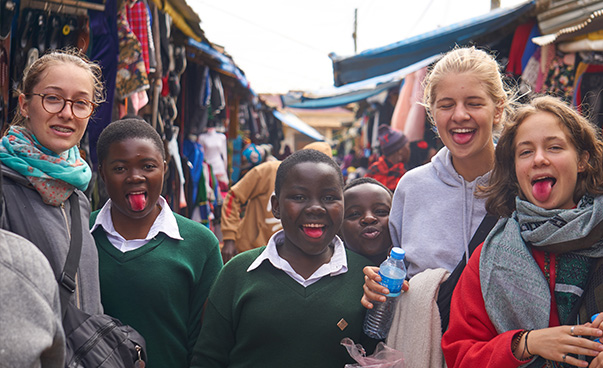 Six schoolgirls stand in a market and smile into the camera. Some stick out a brightly colored tongue.