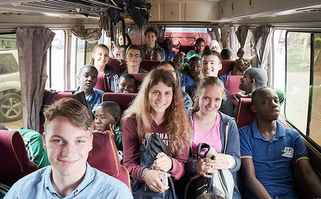 A group of young people sit in a bus and laugh into the camera.