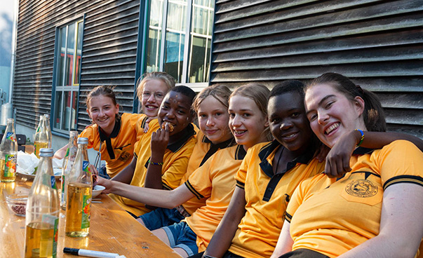 Auf dem Bild sind sieben Schülerinnen zu sehen, die nebeneinander auf einer Bank vor einem Haus sitzen. Alle tragen das gleiche orangefarbene Poloshirt. Sie halten sich teilweise in den Armen. Die Schülerinnen lächeln in die Kamera.