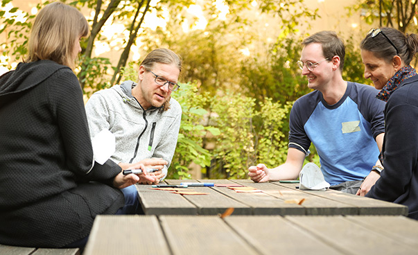 Draußen einem Holztisch sitzen vier Menschen, die sich miteinander unterhalten. In der Mitte des Tisches liegen Moderationskarten, die Person ,die rechts im Bild sitzt hält einen Stift in der Hand.