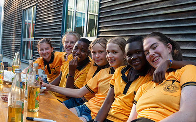 Auf dem Bild sind sieben Schülerinnen zu sehen, die nebeneinander auf einer Bank vor einem Haus sitzen. Alle tragen das gleiche orangefarbene Poloshirt. Sie halten sich teilweise in den Armen. Die Schülerinnen lächeln in die Kamera.