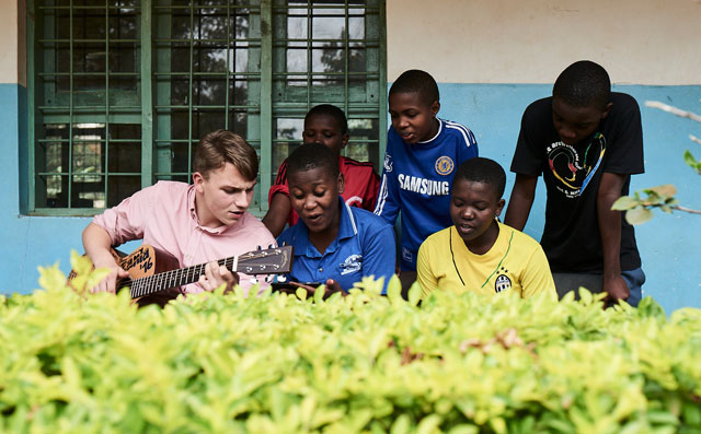 Young people sit together, one plays the guitar and the others sing.