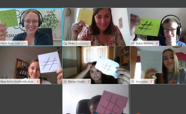 Screenshot von einer Gruppe, deren Portraitaufnahmen in Kachelansicht in einem Video-Chatprogram sichtbar sind und die einen Zettel mit einem # drauf ins Bild halten.