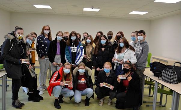 Schülerinnen und Schüler mit Mund-Nasen-Bedeckung stehen bzw. hocken zusammen in einem Klassenzimmer und schauen in die Kamera.