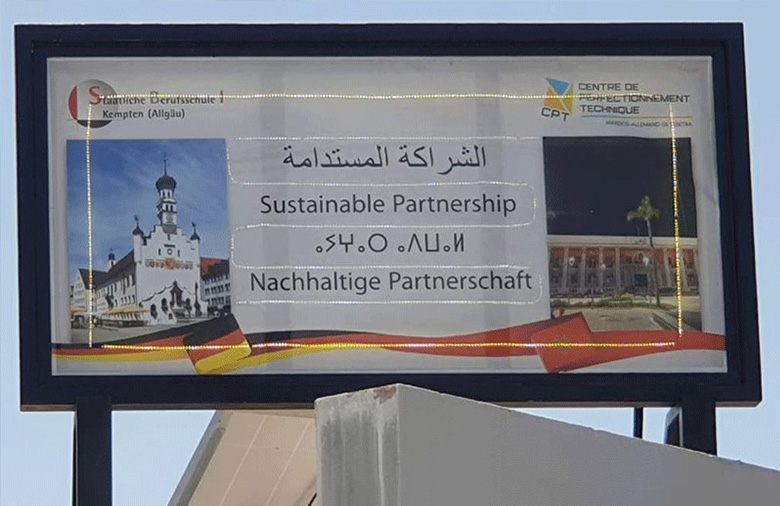 Das fertige Werbeschild auf dem Dach auf dem in vier Sprachen „Nachhaltige Partnerschaft“ steht. Die Logos der beiden Schulen sind in den beiden oberen Ecken zu sehen.