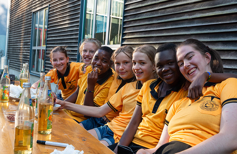 Sieben Schülerinnen sitzen auf einer Bank und lächeln sich in den Armen halten in die Kamera. Alle tragen die gelben Schul T-Shirts der tansanischen Gruppe