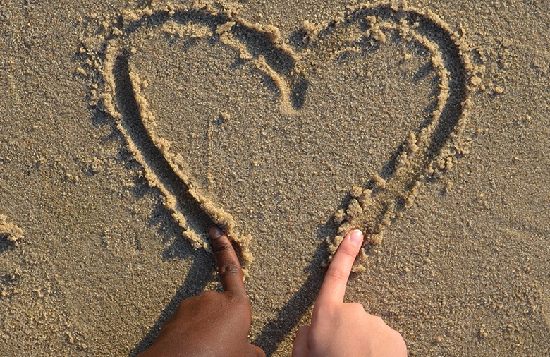 Zwei Menschen zeichnen gemeinsam ein Herz in den Sand. Auf dem Bild sind nur ihre Zeigefinger zu sehen.
