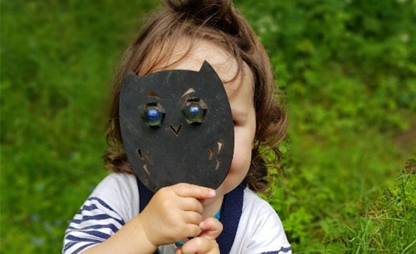 Ein Kind steht in einer Wiese und hät sich eine Eulen-Maske vor das Gesicht.
