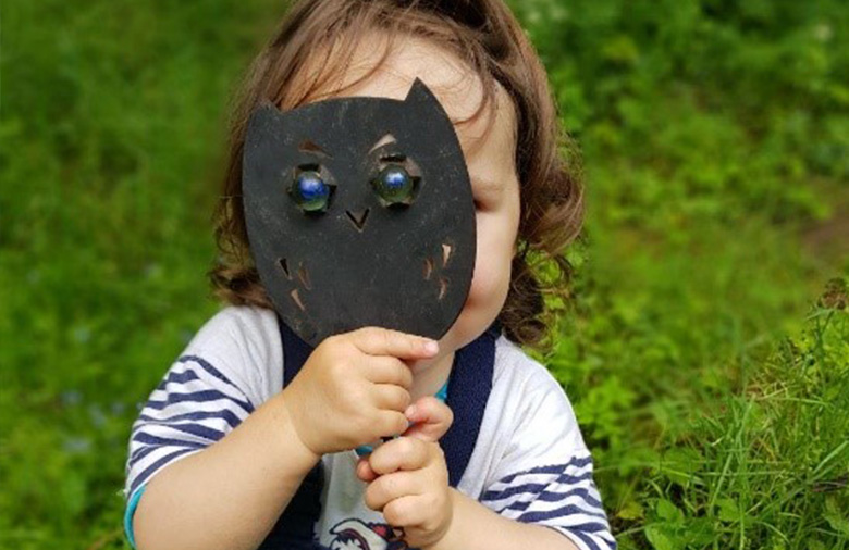 Ein Kind steht in einer Wiese und hät sich eine Eulen-Maske vor das Gesicht.