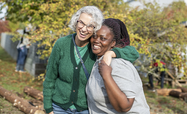 Auf dem Bild sind zwei Frauen zu sehen, die draußen im Grünen stehen und an der Kamera vorbei lächeln. Die Frau auf der linken Seite hat den Arm um die Frau auf der rechten Seite gelegt.