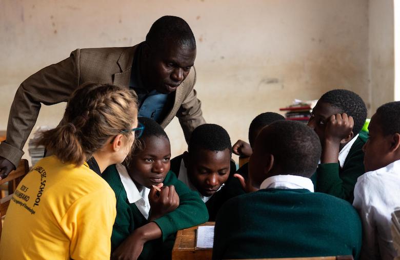 Ein Lehrer schaut einer Gruppe Schülerinnen aus Deutschland und Tansania während der Gruppenarbeit interessiert über die Schulter.